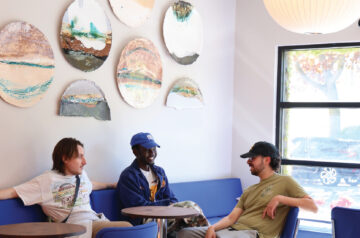 Three people sitting and chatting on a blue cushioned booth in a bright cafe, surrounded by round decorative wall art and modern seating.