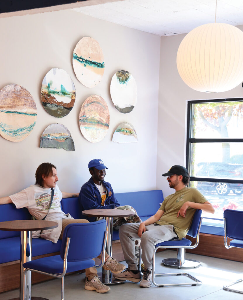 Three people sitting and chatting on a blue cushioned booth in a bright cafe, surrounded by round decorative wall art and modern seating.
