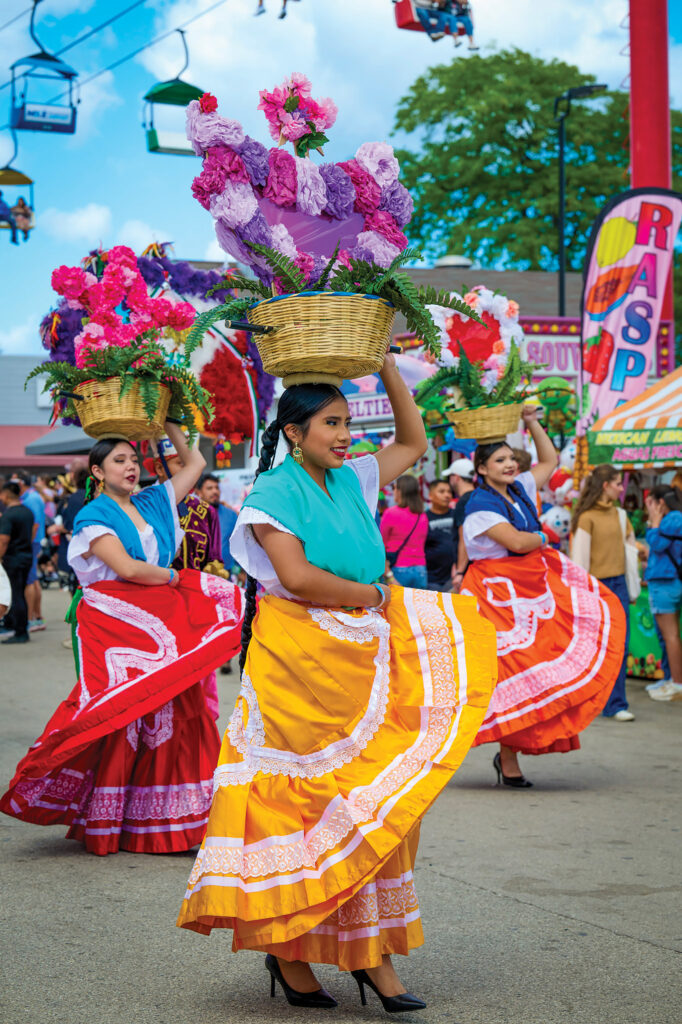 Three women in vibrant Mexican traditional dresses dance in an outdoor festival while balancing woven baskets filled with colorful flowers on their heads.