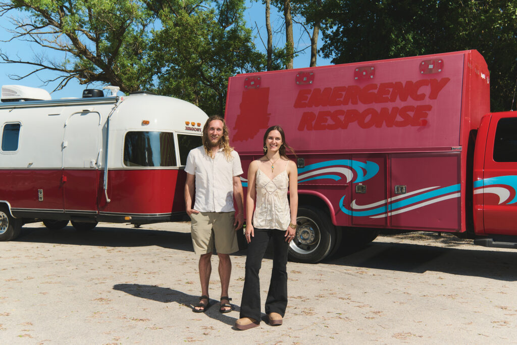 A young couple stands smiling outdoors in front of a red truck attached to a vintage red and white trailer.