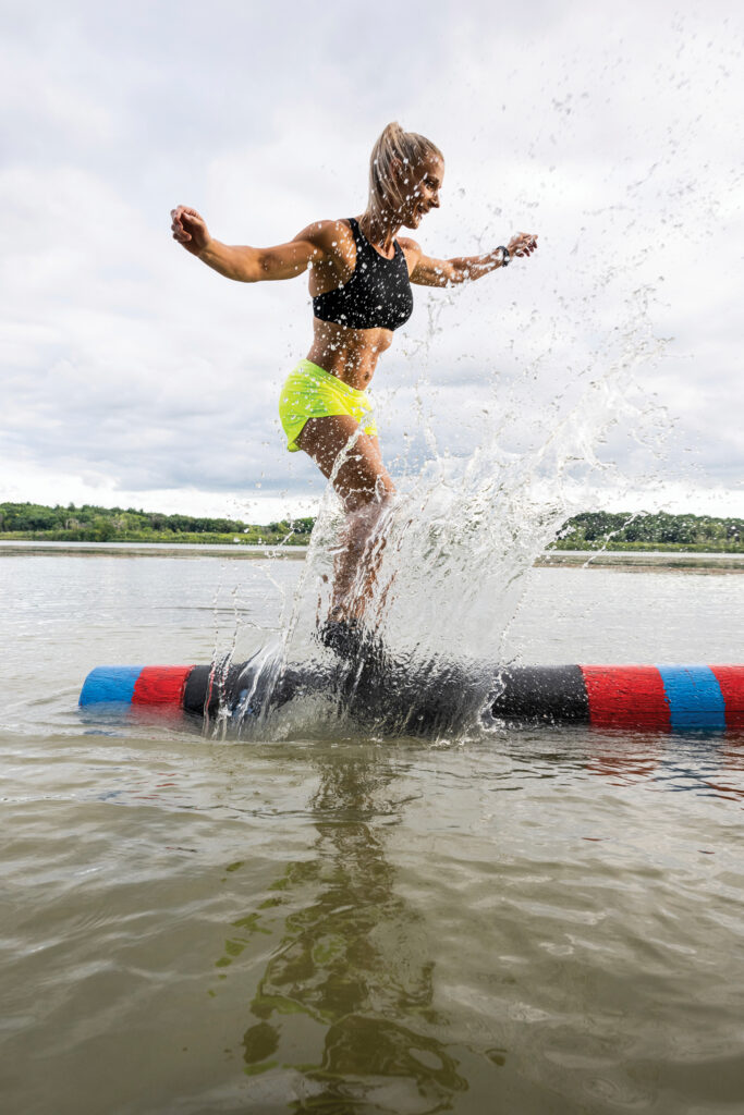 A woman dressed in fitness clothes balances on a log in the water.