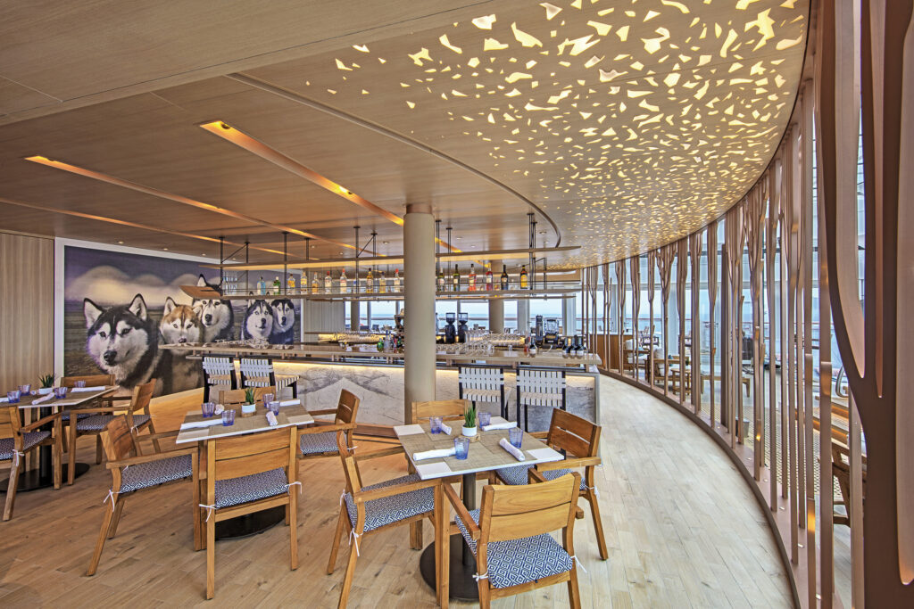 Modern dining area on a cruise ship featuring light wood tables, a glowing mosaic ceiling, bar seating, and a large mural of sled dogs.