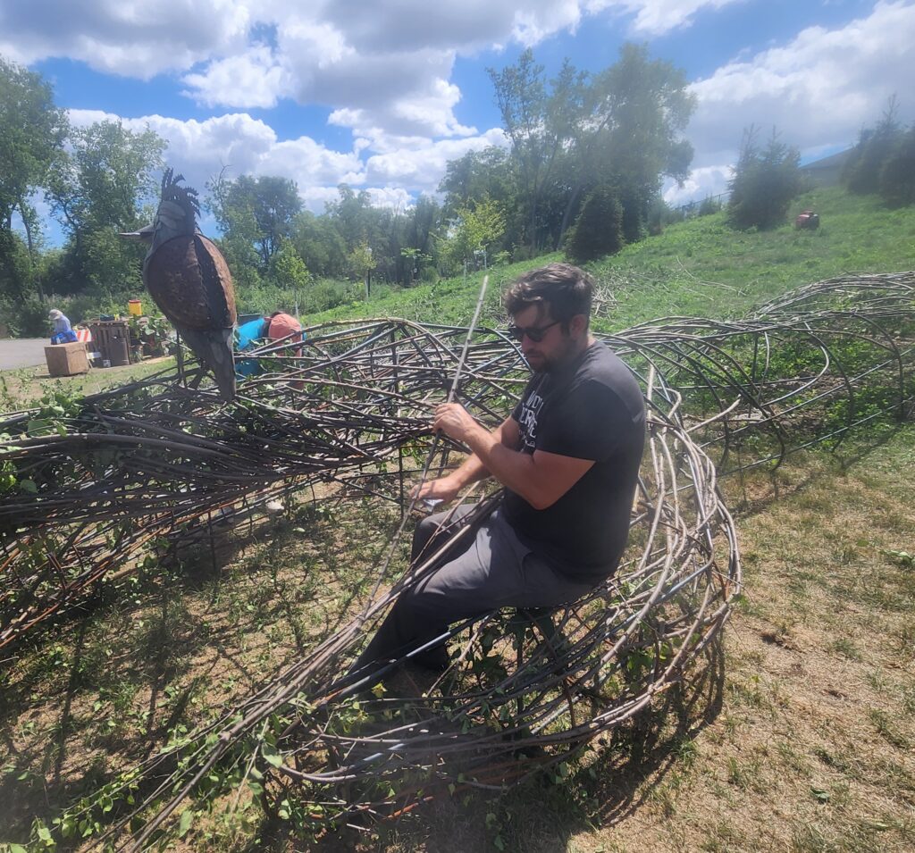 A man is sitting down and weaving with branches in the middle of a field.