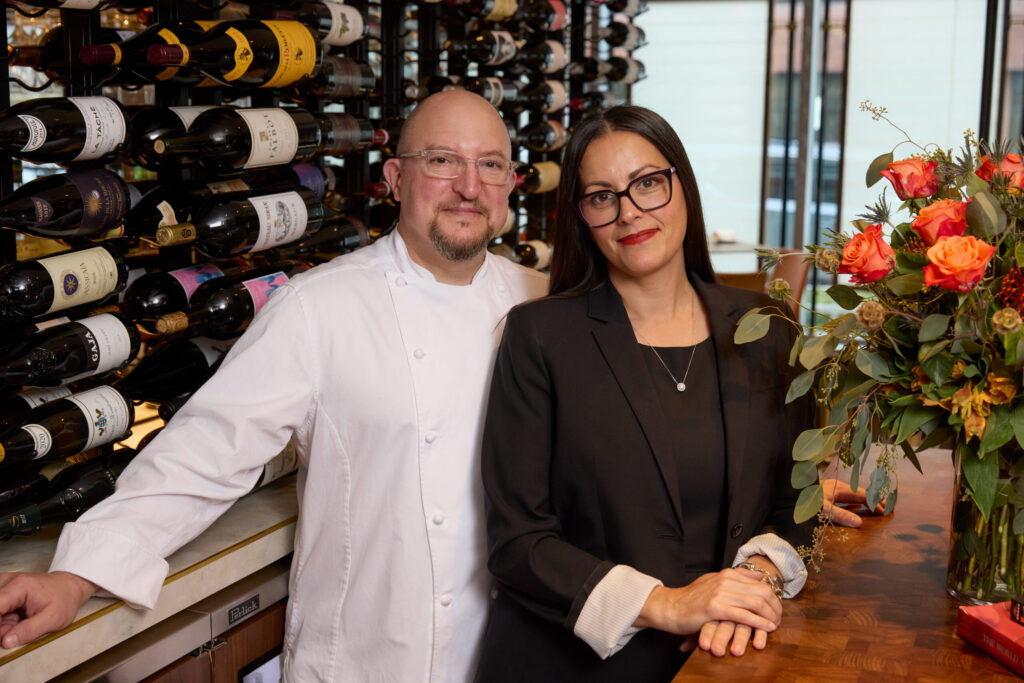 A chef in a white coat and a woman in a black blazer pose together beside an extensive wine rack inside a restaurant, with a large arrangement of orange and coral roses adding color to the scene.