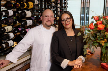A chef in a white coat and a woman in a black blazer pose together beside an extensive wine rack inside a restaurant, with a large arrangement of orange and coral roses adding color to the scene.