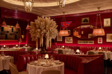 A richly appointed dining room glows in deep crimson and gold, with curved red leather banquettes wrapping around white-tablecloth tables set with china, crystal and small shaded lamps. Oversized arrangements of feathery pampas grass rise dramatically from the center of the room, while pleated pendant shades, brass-framed lanterns and a patterned coffered ceiling add layers of warm, theatrical lighting. The walls are crowded with an eclectic mix of framed art, textiles and decorative objects, including a large geometric tapestry and a faceted, gold-edged mirror. Soft upholstered dining chairs, polished dark floors and carefully arranged place settings reinforce the room’s formal, clubby atmosphere.