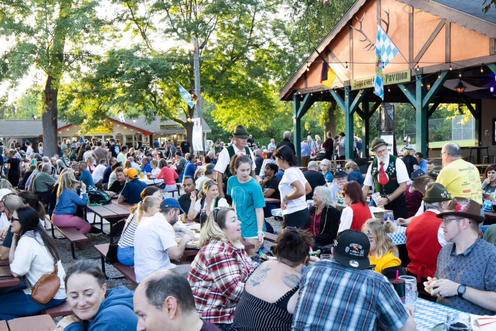 A lively crowd fills picnic tables at an outdoor German-style beer garden, where guests eat, drink and socialize beneath mature shade trees and strings of lights. Servers in traditional German-inspired attire move among the tables near the open-air Jägermeister Pavilion, which is decorated with German and Bavarian flags, adding to the festive beer-hall atmosphere.