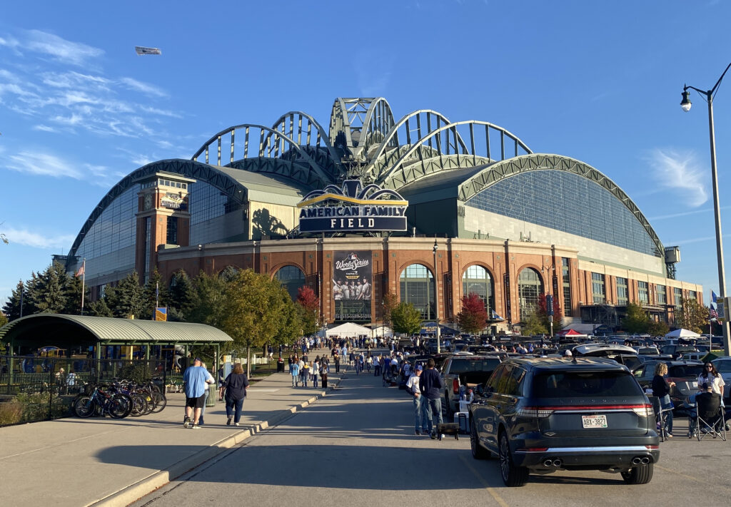 A crowd of Milwaukee Brewers fans gathers outside American Family Field on a sunny day, with cars filling the parking lots and people walking toward the stadium. A large World Series banner hangs above the main entrance, framed by the ballpark’s distinctive arched roof and brick facade.