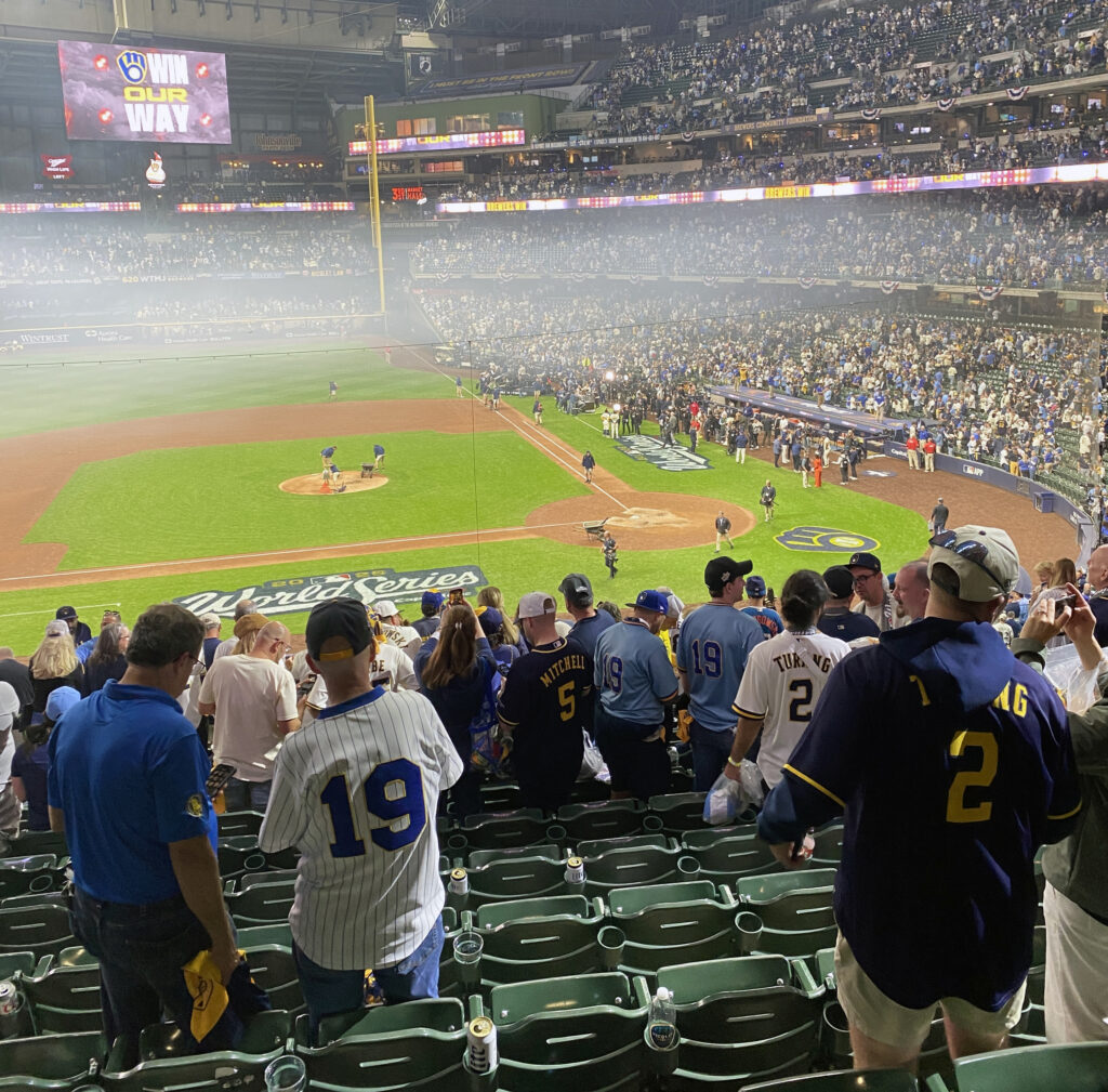 Fans in Milwaukee Brewers jerseys begin leaving their seats at American Family Field following a World Series game. Grounds crew members work on the pitcher’s mound as a haze hangs over the field, while crowds remain in the stands and the scoreboard displays the team’s “Win Our Way” slogan.