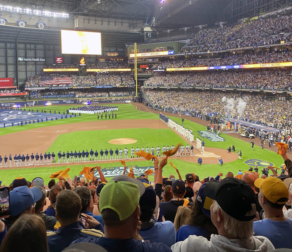 Milwaukee Brewers and Tampa Bay Rays players line the baselines during pregame ceremonies for the World Series at American Family Field. A massive American flag stretches across the outfield, smoke rises near the dugout and thousands of fans fill the stands, with spectators in the foreground waving gold rally towels.