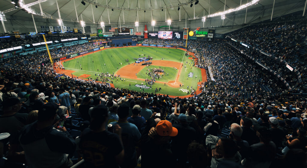 A panoramic view of Tropicana Field in St. Petersburg, Florida, shows fans filling the stands as players, media members and other personnel gather on the field following a World Series game. The stadium’s video boards display “Thank You Fans” alongside the Tampa Bay Rays logo.