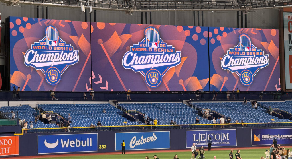 Three massive video screens at Tropicana Field display graphics celebrating the Milwaukee Brewers as 2026 World Series champions. The colorful blue, purple and orange designs feature the Brewers’ glove logo beneath the words “World Series Champions,” while fans and stadium personnel linger among the mostly empty blue seats below.