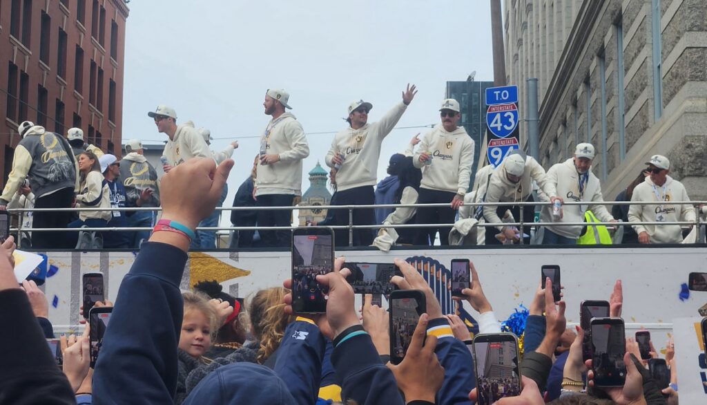 Milwaukee Brewers players celebrate aboard an open-top parade bus, wearing cream-colored World Series championship hoodies and caps as they wave to fans lining a downtown Milwaukee street. Spectators in the foreground raise their phones to capture the moment, with historic buildings and interstate signs in the background.