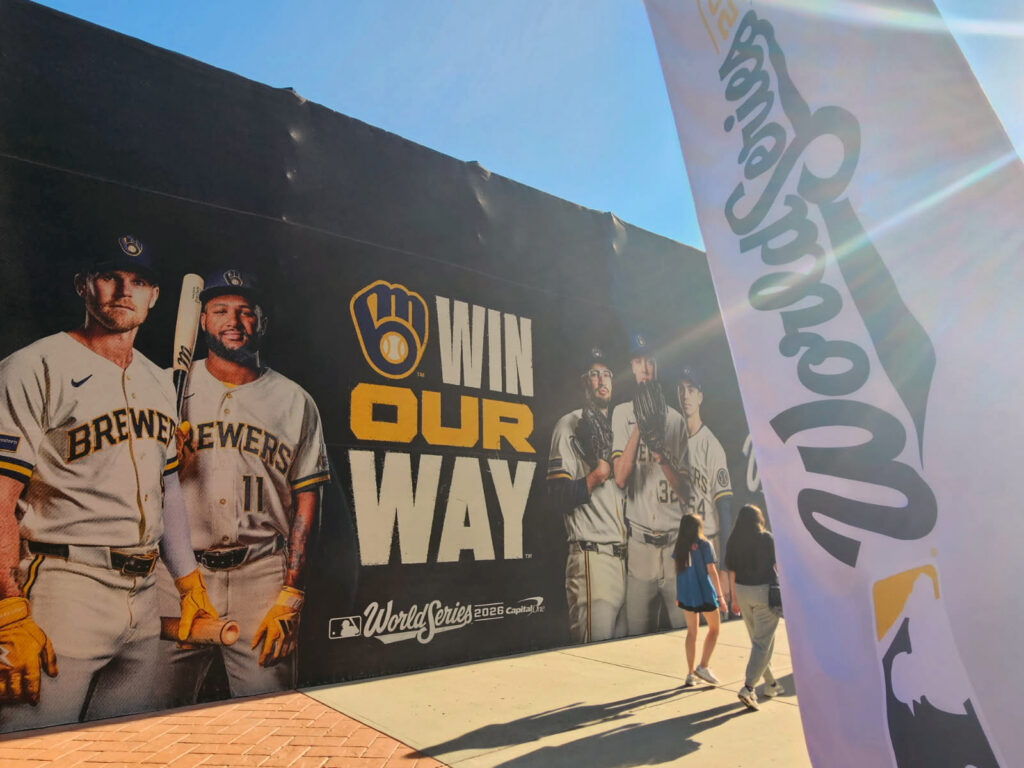 A massive Milwaukee Brewers promotional mural outside American Family Field features images of players in uniform alongside the slogan “WIN OUR WAY” and the 2026 World Series logo. Two people walk past the display in bright sunlight, while a large white Brewers flag dominates the right side of the image.