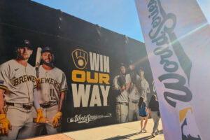 A massive Milwaukee Brewers promotional mural outside American Family Field features images of players in uniform alongside the slogan “WIN OUR WAY” and the 2026 World Series logo. Two people walk past the display in bright sunlight, while a large white Brewers flag dominates the right side of the image.